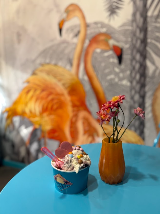 A cup of gelato set beside a small vase of flowers with a flamingo graffiti in the background wall